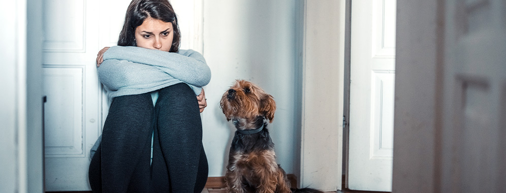 Eine Frau mit müdem Gesichtsausdruck und verschränkten Armen, sitzt neben einem Hund auf dem Boden.